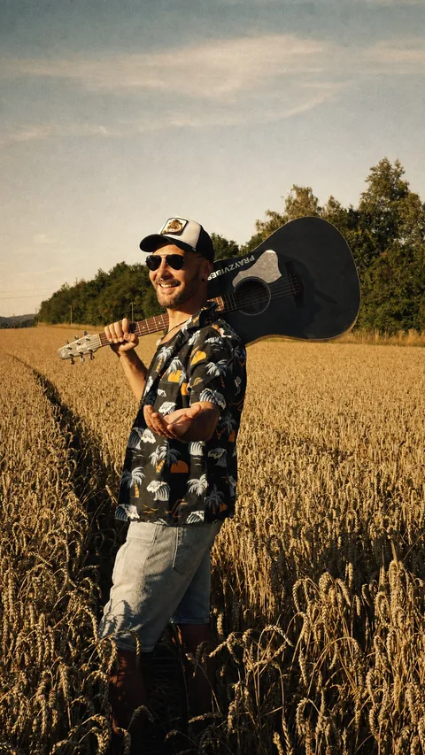 PRAYZ smiling in a field with a black acoustic guitar over one shoulder