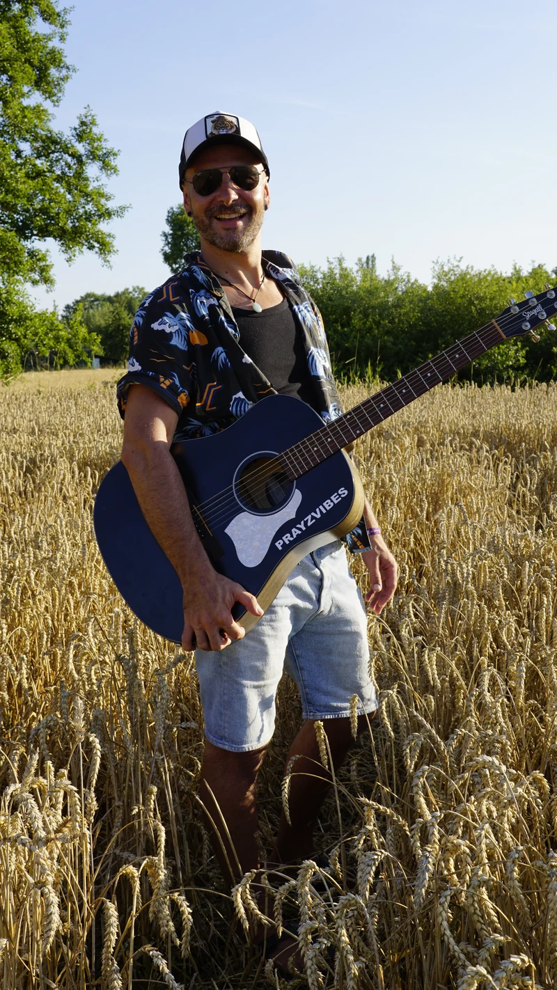 PRAYZ with a black acoustic guitar in a cornfield
