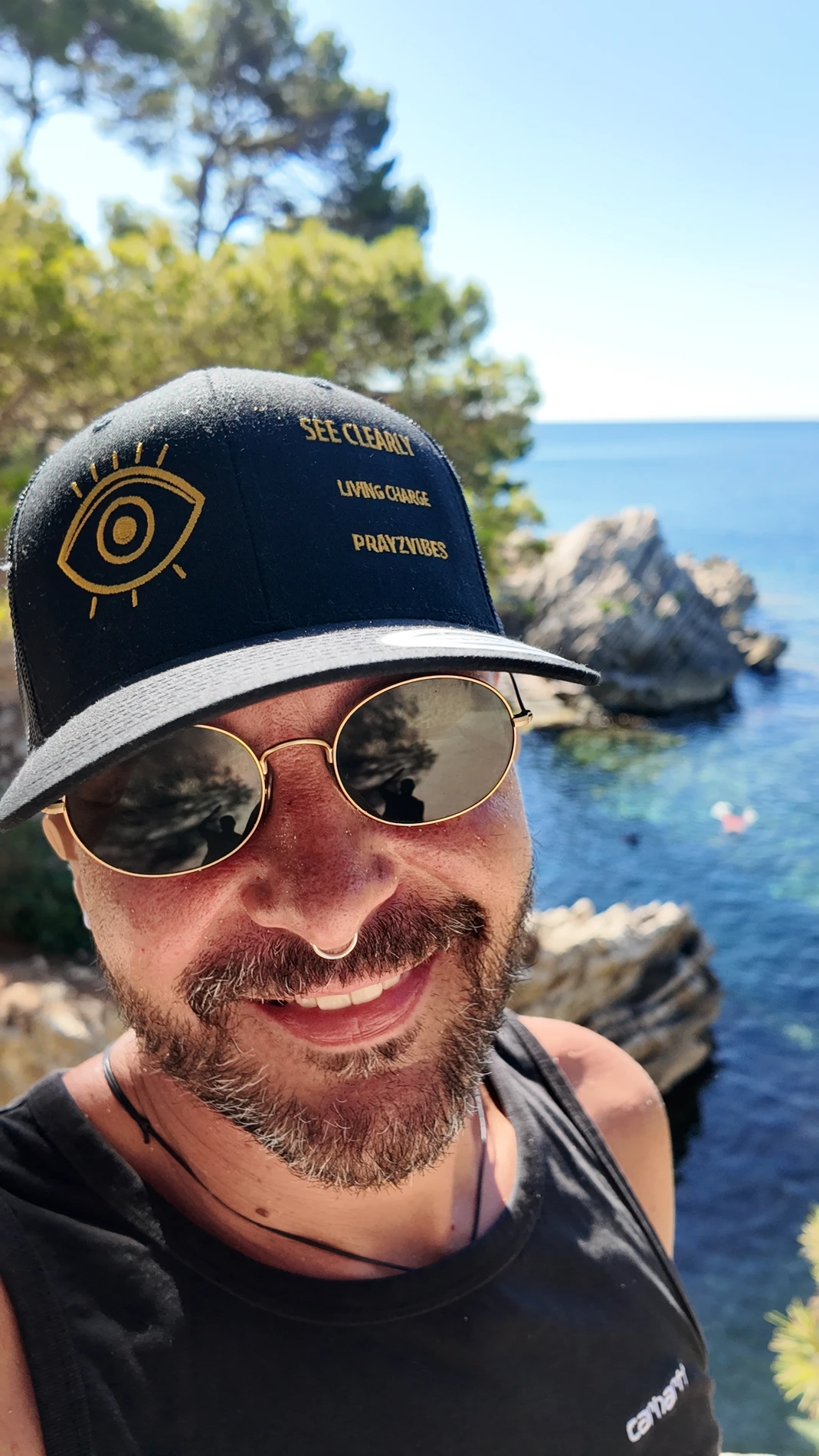 PRAYZ smiling by the sea, wearing his black SEE CLEARLY cap with gold embroidery